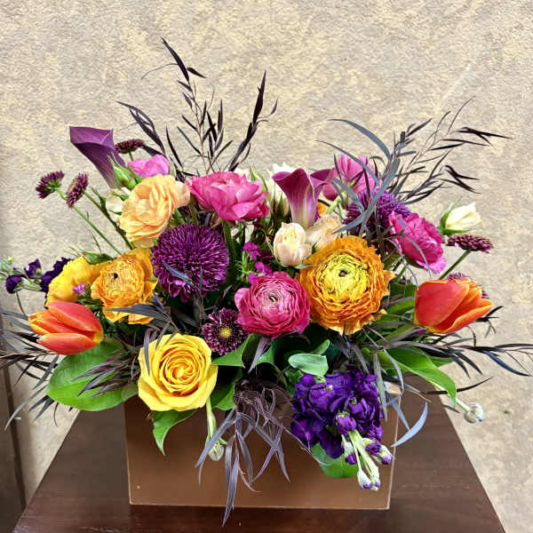 Low rectangular arrangement of bright yellow, orange, pink, and purple flowers in a brown box.