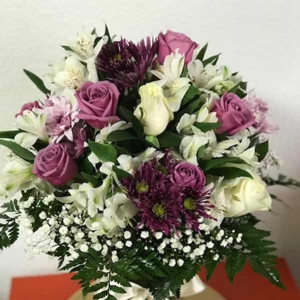 Round arrangement of purple and white flowers in a clear vase with white ribbon on a gold plate
