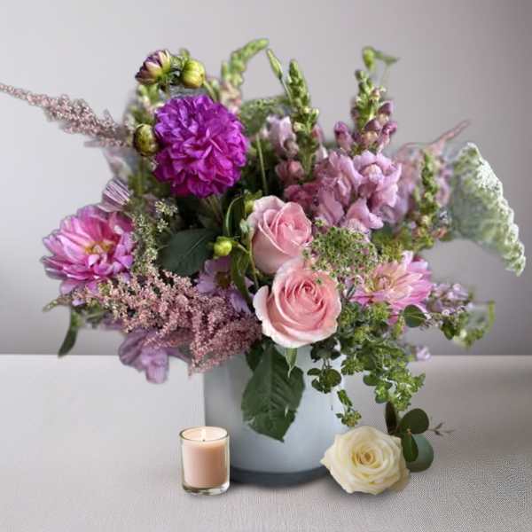 Pink and purple flower arrangement in a white vase with a candle and rose