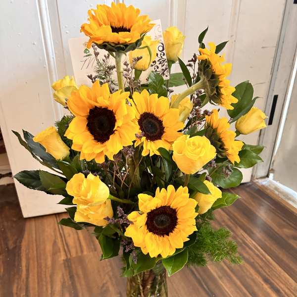 Tall vase arrangement of yellow sunflowers and yellow roses with a small card.