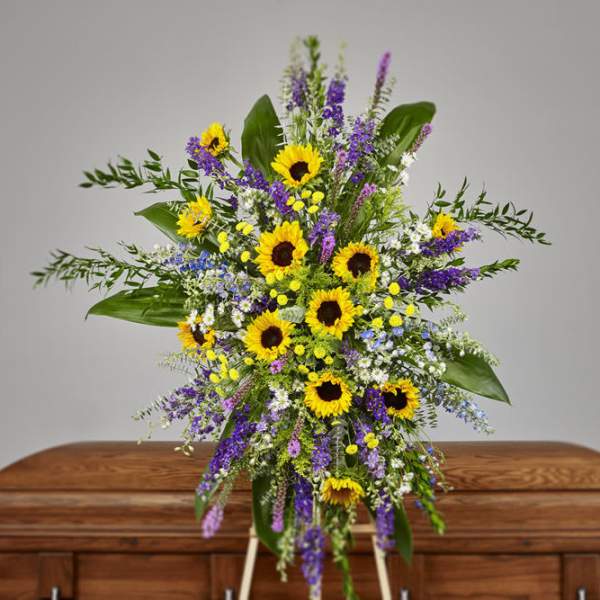 Standing spray of yellow sunflowers and purple flowers in front of a wooden casket