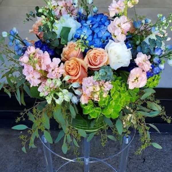 Large arrangement of blue hydrangeas, peach roses, and mixed blooms in a vase on a clear stand