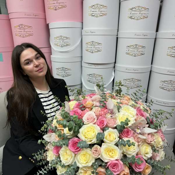 Large pink hatbox arrangement of pastel roses with orchids, next to a seated woman.