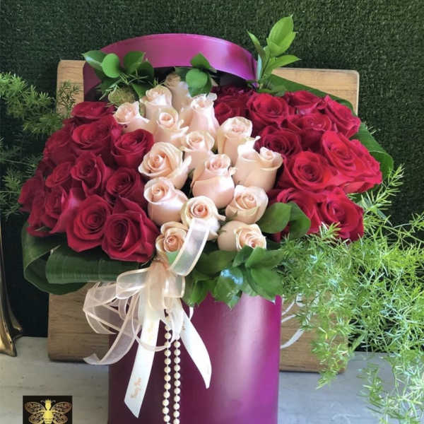 Round magenta hatbox filled with red and blush roses, trailing greenery, and a ribbon bow.