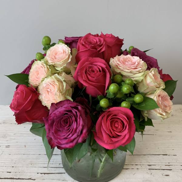 Low arrangement of bright pink and blush roses with green berries in a clear glass vase