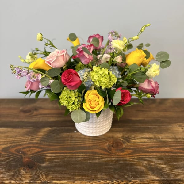 Low arrangement of mixed roses and other blooms in pink, yellow, and red in a white vase