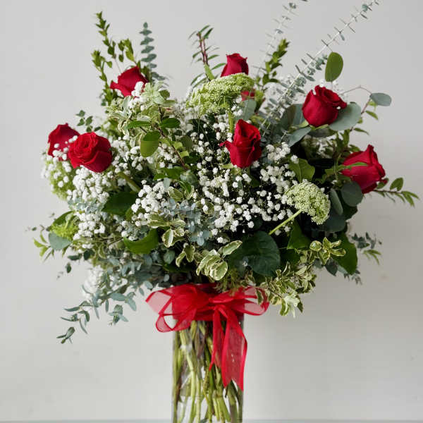 Tall bouquet of red roses, white filler flowers, and greenery in a clear glass vase with a red ribbon.