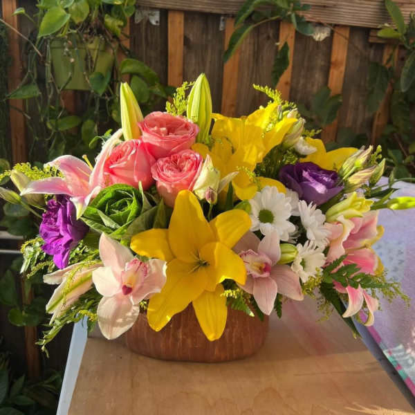 Mixed bouquet with pink roses, yellow lilies, and purple blooms in a wooden bowl