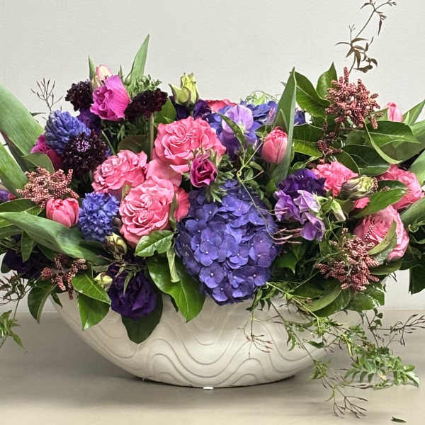Low oval arrangement of pink roses and tulips with purple hydrangeas in a white ceramic bowl