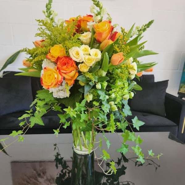 Tall vase of yellow and orange roses with white blooms and trailing ivy