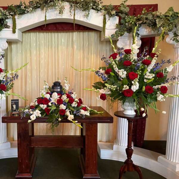 Symmetrical funeral floral display with red and white arrangements on pedestals and a table centerpiece