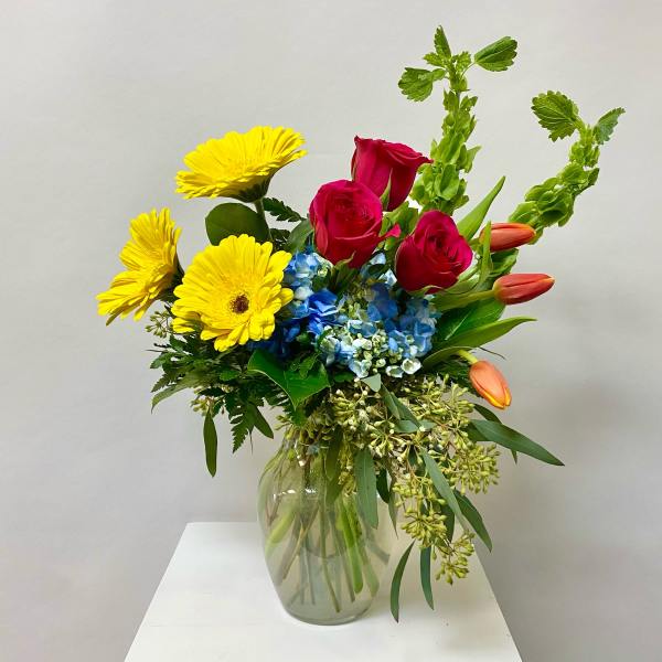 Bouquet of yellow daisies, red roses, blue hydrangea, and orange tulips in a glass vase
