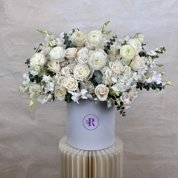 Large white and cream rose and ranunculus arrangement in a round white hatbox on a pleated pedestal.
