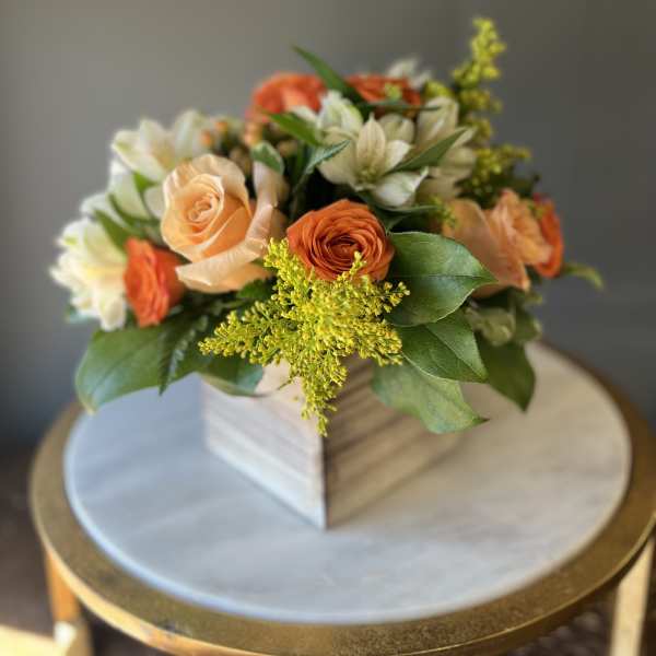 Compact arrangement of orange and peach roses with white blooms in a wooden cube vase.