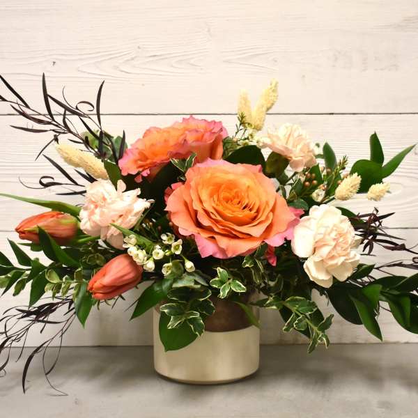 Low arrangement of peach roses, blush carnations, and orange tulips in a cream ceramic vase