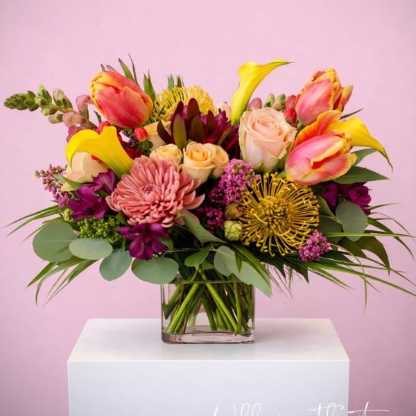 Colorful arrangement of yellow calla lilies, roses, tulips, and protea in a clear glass cube vase