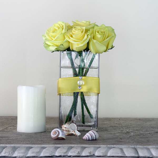 Yellow roses in a clear rectangular vase with a yellow ribbon and candle beside it