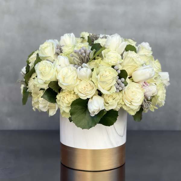 Low arrangement of cream roses and white tulips in a white and gold cylinder vase