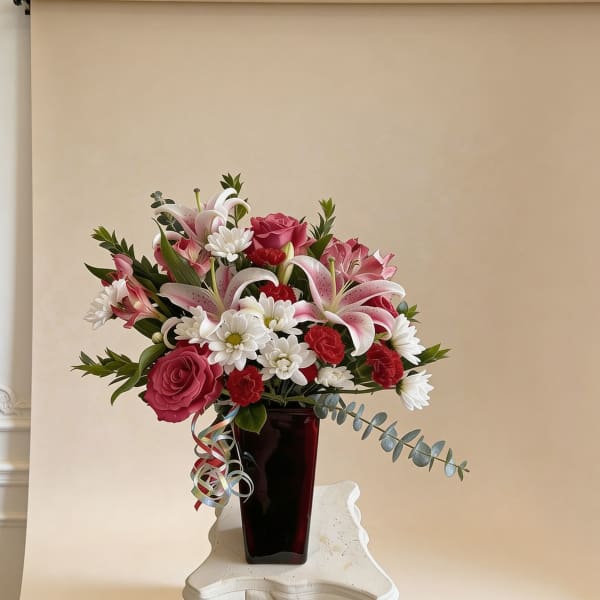 Arrangement of pink lilies, red roses, red carnations, and white daisies in a dark red vase