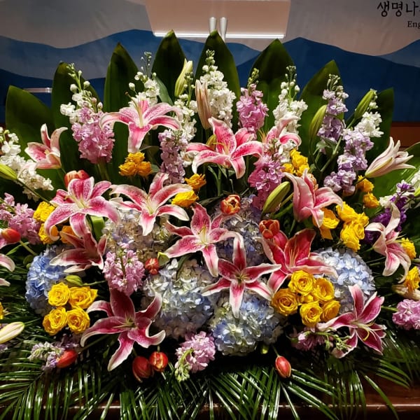 Large fan-shaped arrangement of pink lilies, yellow roses, blue hydrangeas and pastel blooms framed by glossy green leaves