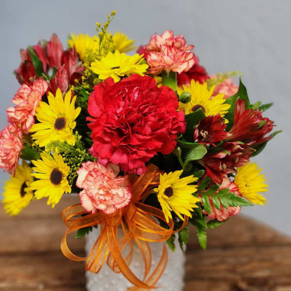 Bright arrangement of red and pink carnations with yellow daisies in a white vase with orange ribbon