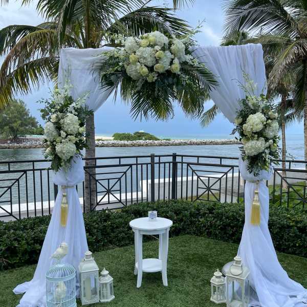 White floral wedding arch with draped fabric and lanterns by the water