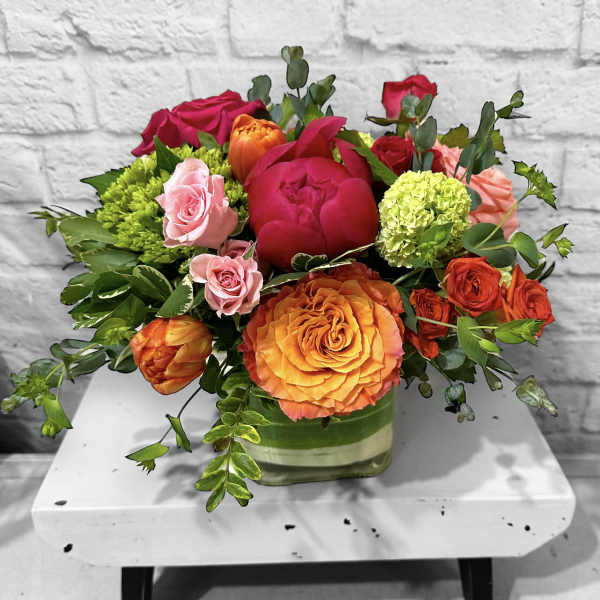 Low arrangement of orange and pink roses, peonies, and tulips in a square glass vase