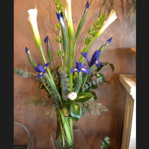 Tall arrangement of white calla lilies, blue irises, and green bells of Ireland in a clear glass vase.