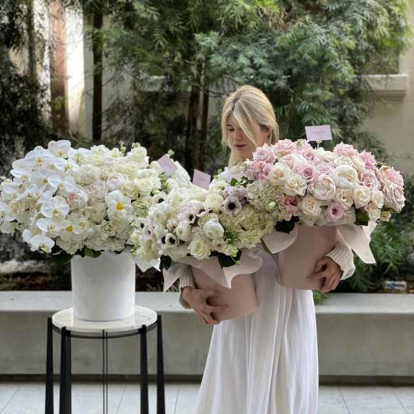 Woman holding two large pastel flower bouquets beside a white floral arrangement.