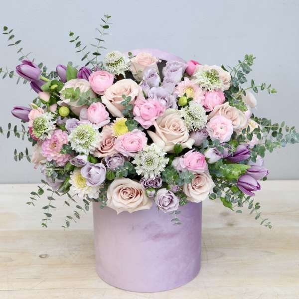 Pastel pink and lavender flower arrangement in a round lavender hatbox-style container