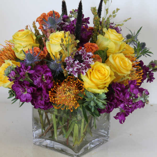 Bouquet of yellow roses and purple flowers in a clear square vase