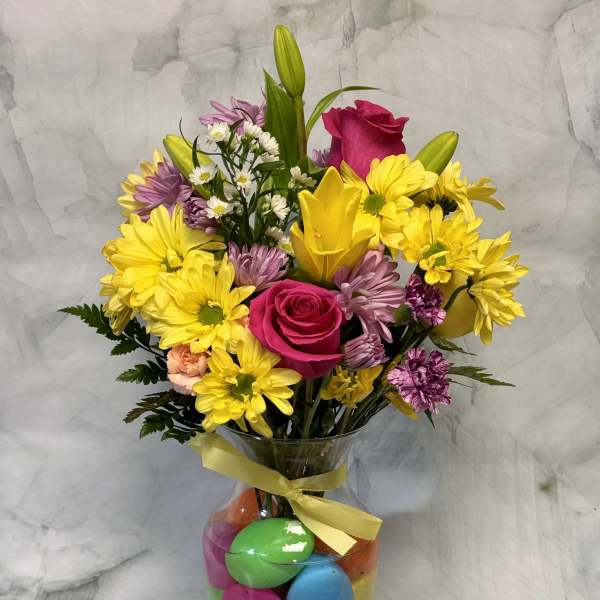 Bouquet of yellow daisies, pink roses, and lilies in a glass vase with colorful eggs