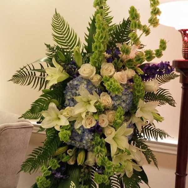 Tall standing spray of white lilies, roses, blue hydrangeas, and green bells of Ireland on an easel