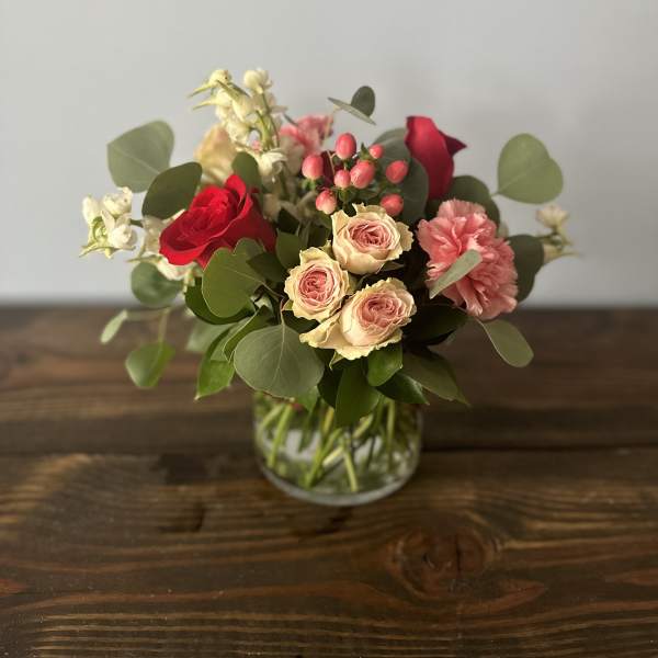 Short arrangement of red roses, blush spray roses, pink carnations, and white flowers in a clear glass vase