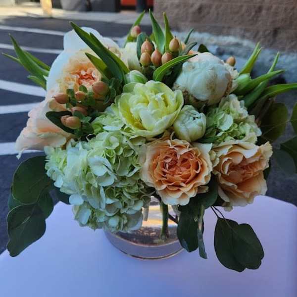 Low arrangement of peach and cream flowers with green hydrangeas in a round metallic vase.