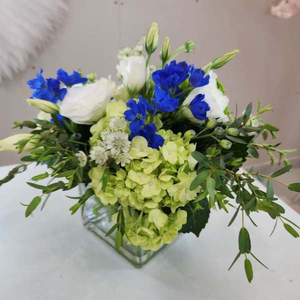 Low arrangement of green hydrangeas, blue delphinium, and white blooms in a clear glass cube vase