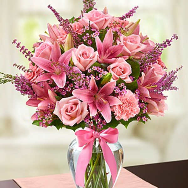 Pink lilies, roses, and carnations arranged in a clear glass vase with a pink bow