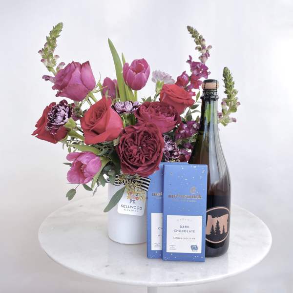 Bouquet of pink and red flowers in a white vase with chocolate bars and a bottle