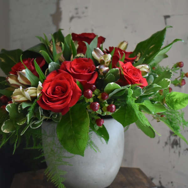 Red roses arranged in a white vase with green foliage and berry accents