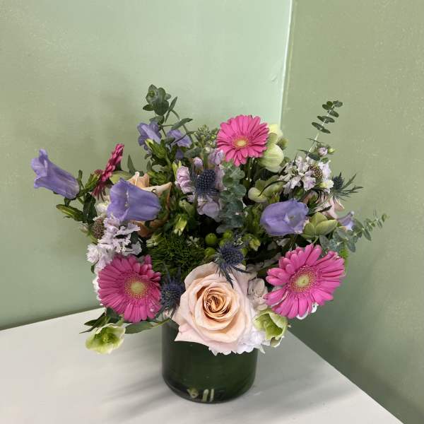Mixed arrangement of pink gerbera daisies, peach roses, and purple blooms in a green glass cylinder vase