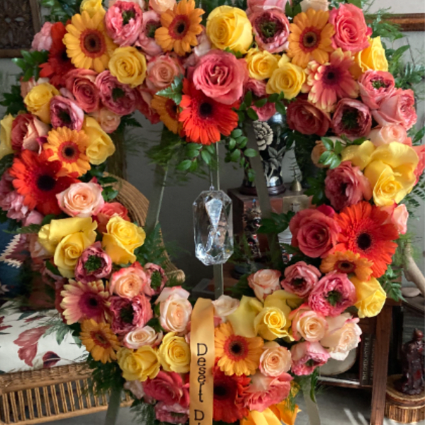 Heart-shaped standing wreath of pink, yellow, and orange roses and gerbera daisies on an easel with ribbon.