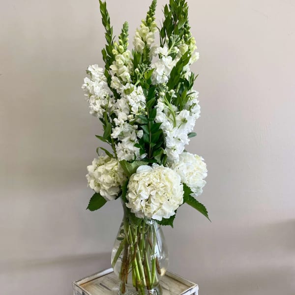 Tall white hydrangeas and snapdragons arranged in a clear glass vase on a small wooden stand