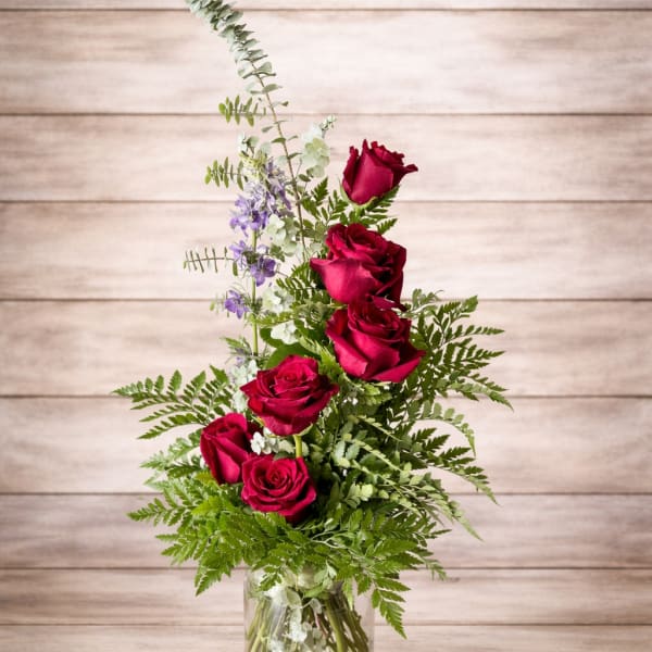Red roses in a clear glass vase with mixed greenery and small purple flowers