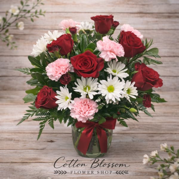 Red roses, pink carnations, and white daisies in a glass vase with a ribbon