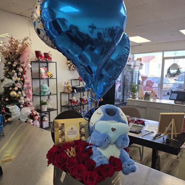 Heart-shaped blue balloons above red roses, chocolates, and a blue plush toy in a box
