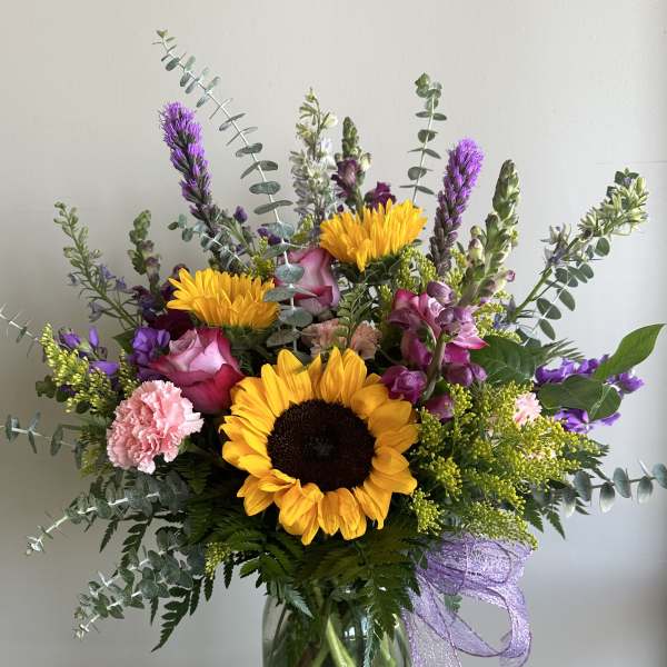 Tall mixed arrangement with sunflowers, pink roses, and purple flowers in a clear glass vase