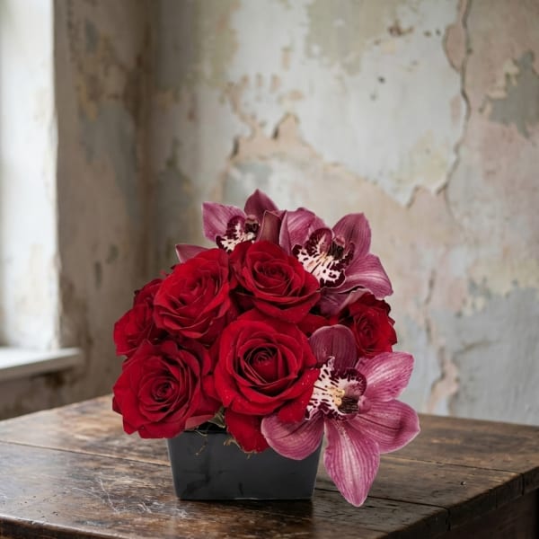 Low arrangement of red roses and mauve orchids in a black square vase on a rustic wooden table.