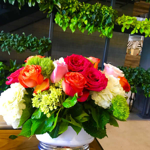 Low arrangement of red, orange, and pink roses with white hydrangeas in a white bowl vase