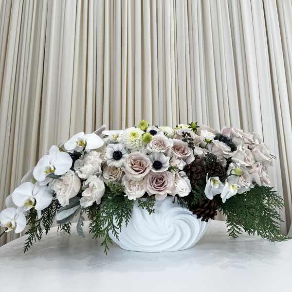 Low centerpiece of white orchids, blush roses, anemones, and evergreens in a white ceramic bowl