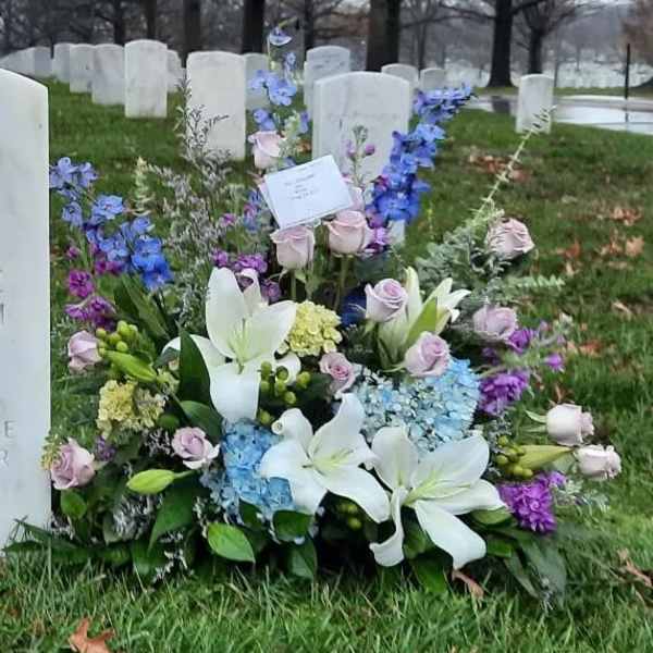 Sympathy arrangement of white lilies, pastel roses, and blue flowers placed at a cemetery headstone.